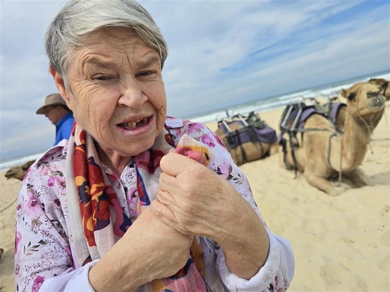 An older disabled lady stands in front of camels at the beach.